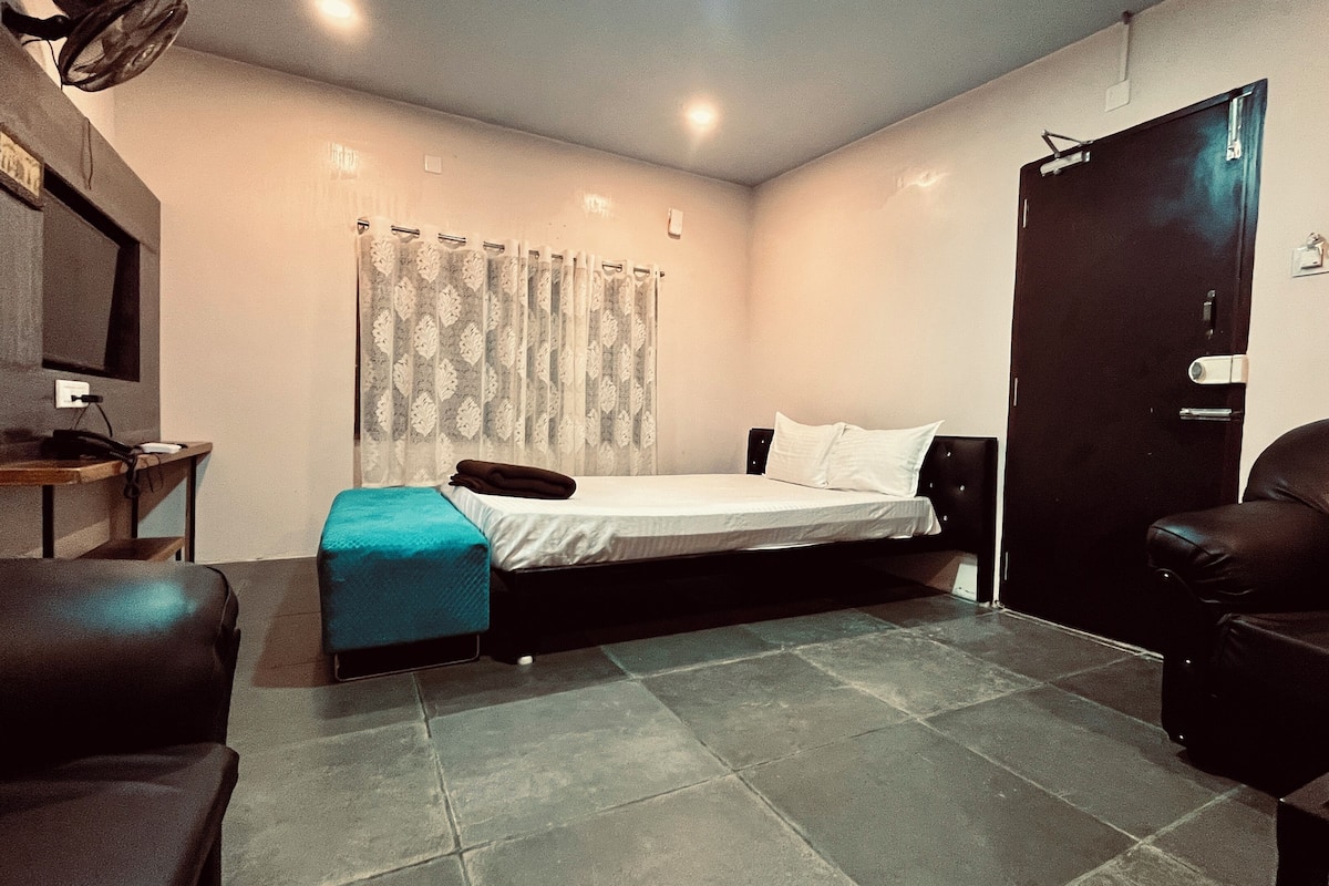 A small, well-lit room with a bed, a desk, a chair, and a door.