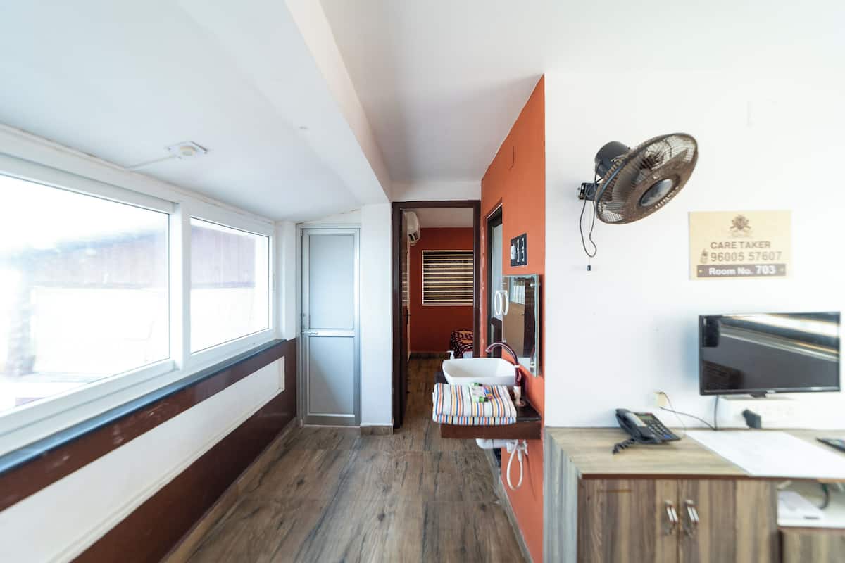 A hotel room with a wooden floor, a window, a sink, a mirror, a fan, a desk with a computer, and a telephone.