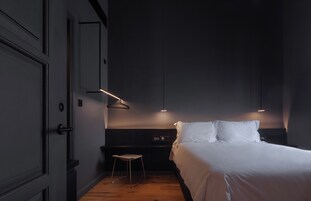 Quarto de hotel moderno com uma cama grande, um banquinho pequeno e duas luminárias pendentes.