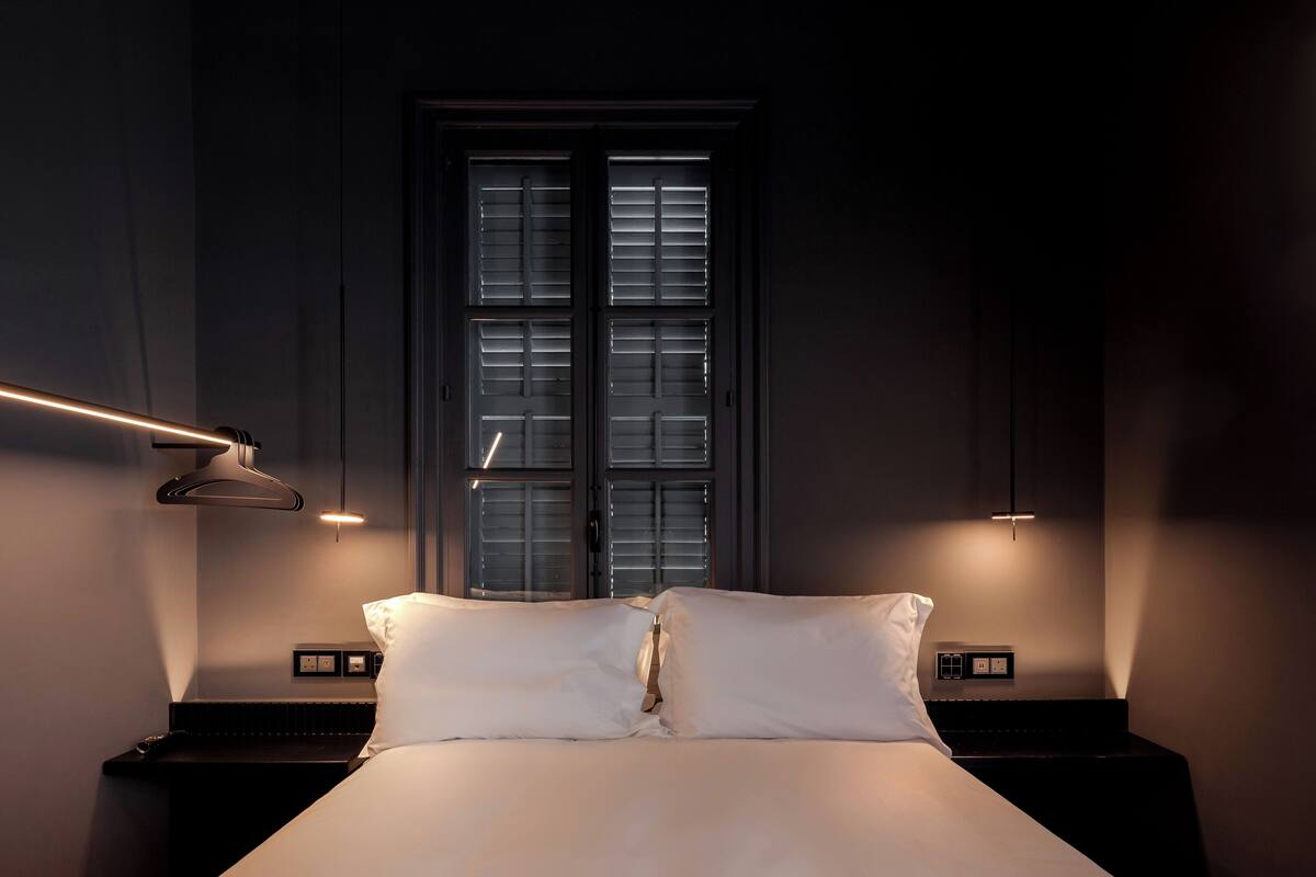 Ein ordentlich bezogenes Bett mit weißen Leinen, ein Nachttisch mit Lampe und ein Fenster mit Jalousien.