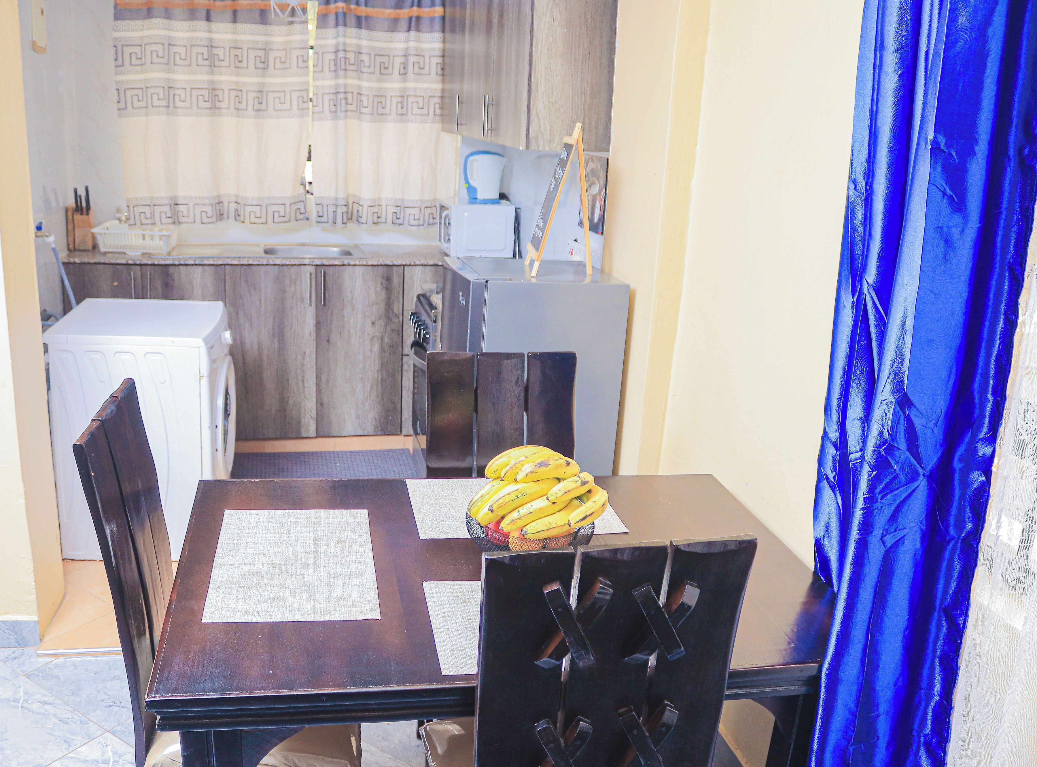 Un coin repas avec une table, des chaises, un panier de bananes et, au fond, une cuisine.