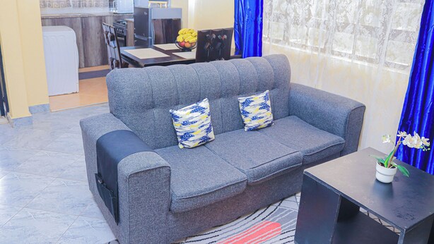 A grey sofa with patterned cushions, a small table with a potted plant, and a kitchen area in the background. - Countryside Apartments -With Unlimited WIFI&Netflix (Nairobi)