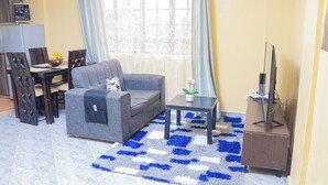 A living room with a sofa, a coffee table, a TV stand, and a dining area with chairs and a table. - Countryside Apartments -With Unlimited WIFI&Netflix (Nairobi)