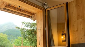 A bedroom with a large window, wooden ceiling beams, a bed with a grey mattress, and a wall-mounted light fixture. - Superbe Appartement Chalet Pieds des Pistes et des Bains (Le Monêtier-les-Bains)