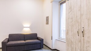 A modern room with a dark grey sofa, a wooden wardrobe, a wall-mounted light, and a window with blinds. - Studios 3 in Napoli With 1 Bathrooms (Naples)
