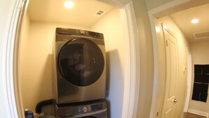Une buanderie équipée de machines à laver et de sèche-linge superposés, d’une poubelle et d’une porte donnant sur une autre pièce.