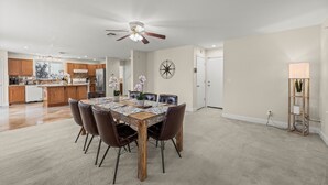 Un grand espace à manger avec une table et des chaises en bois, un ventilateur de plafond et une cuisine en arrière-plan.