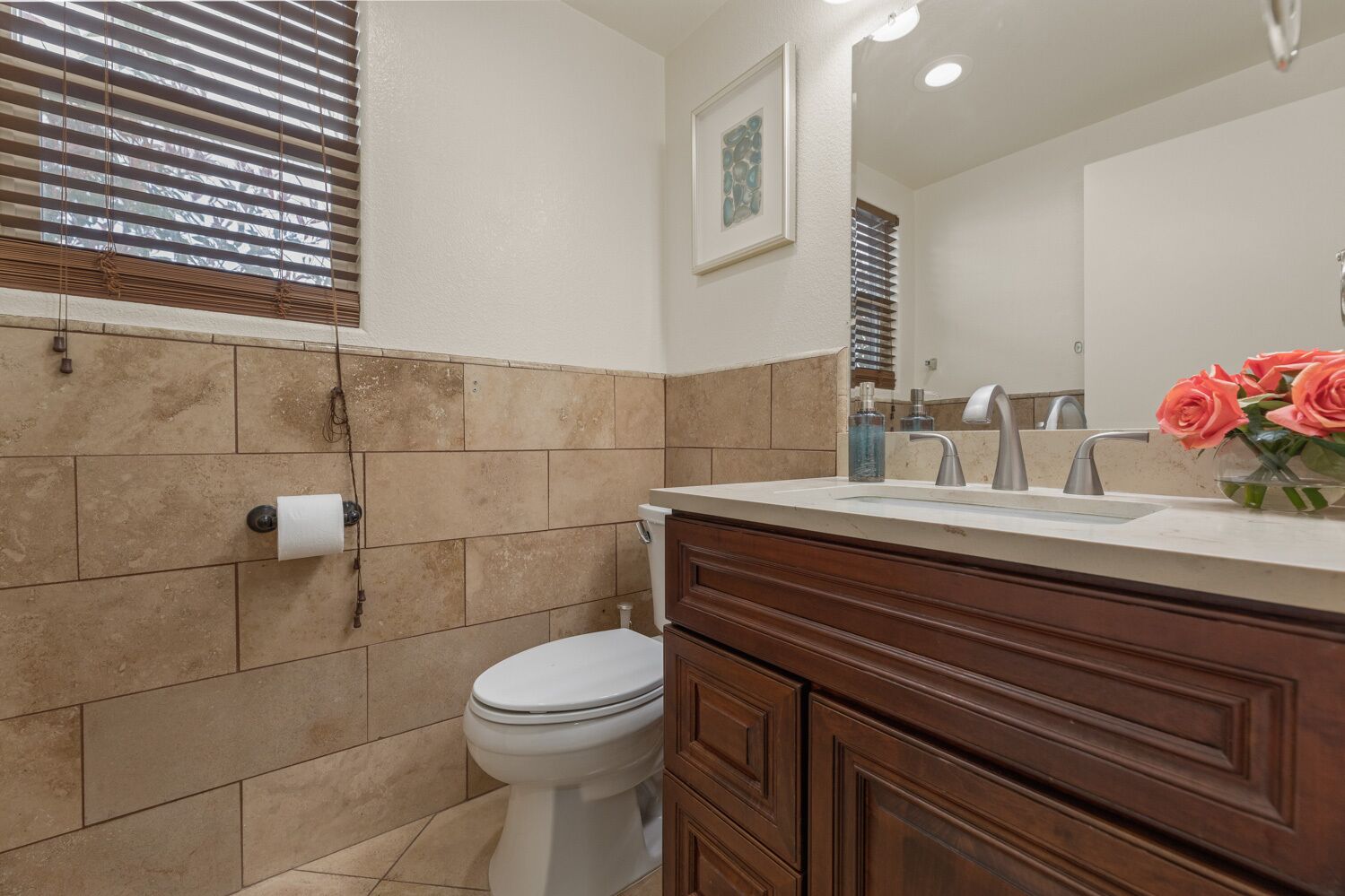 A bathroom with beige tiles, a white toilet, a wooden vanity with a sink, and a framed picture on the wall.