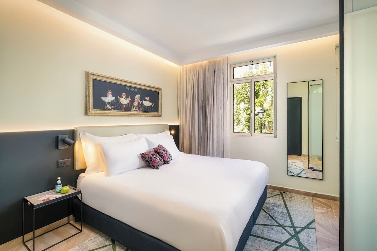 Une chambre d’hôtel avec un grand lit, une table de chevet sur laquelle se trouvent une bouteille et une pomme, un tableau encadré au mur, une fenêtre avec des rideaux et un miroir.