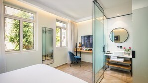 A modern bedroom with a large bed, a desk with a computer, a mirror, and a window with a view of trees. - Rephael House Boutique Hotel (Rishon LeTsiyon)