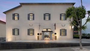 A large, multi-story building with a symmetrical facade, featuring evenly spaced windows with shutters, a central entrance with a decorative arch, and a small tree in the foreground. - Rephael House Boutique Hotel (Rishon LeTsiyon)