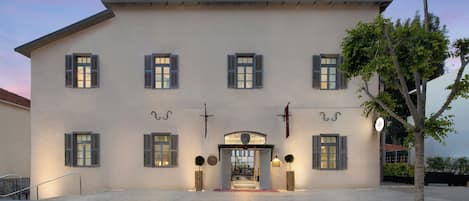 A large, multi-story building with a symmetrical facade, featuring evenly spaced windows with shutters, a central entrance with a decorative arch, and a small tree in the foreground.