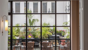 A view through a large window with multiple panes, revealing an outdoor seating area with wicker chairs and tables, surrounded by lush greenery and potted plants. - Rephael House Boutique Hotel (Rishon LeTsiyon)