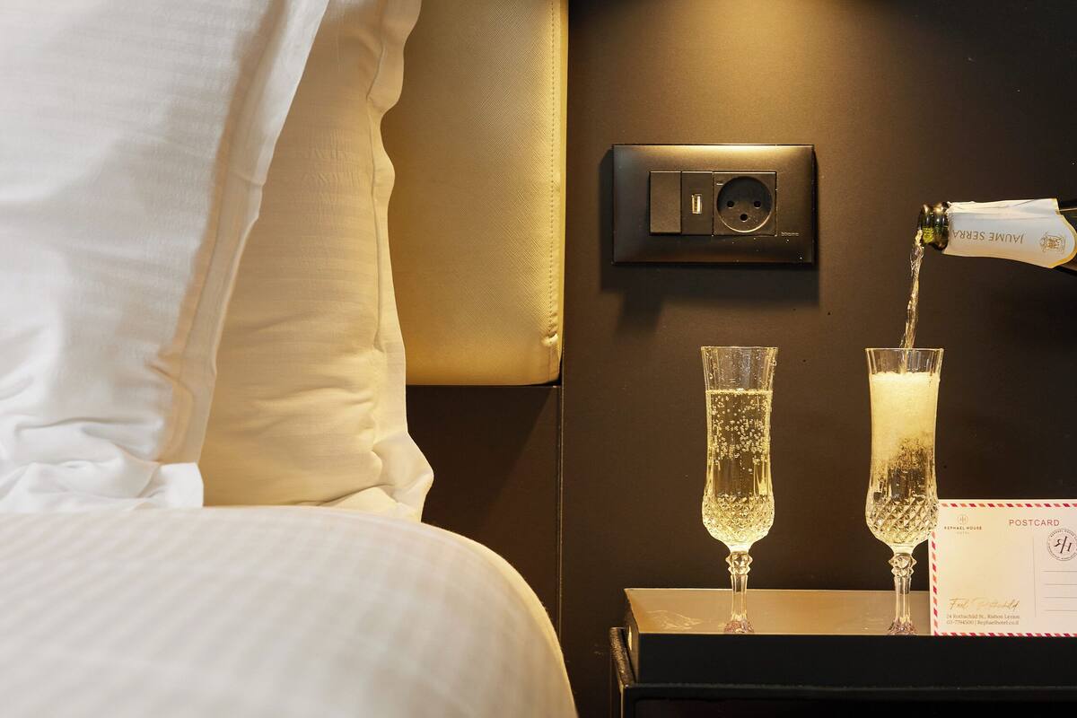 Une chambre d’hôtel avec un lit parfaitement fait, un luminaire fixé au mur et deux coupes de champagne posées sur une table de chevet.