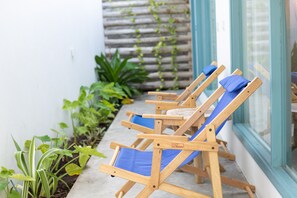 Une rangée de chaises longues en bois, dotées de coussins bleus, est disposée le long d’un chemin de béton, à côté d’un bâtiment avec une porte en verre et une clôture en bois recouverte de plantes grimpantes.
