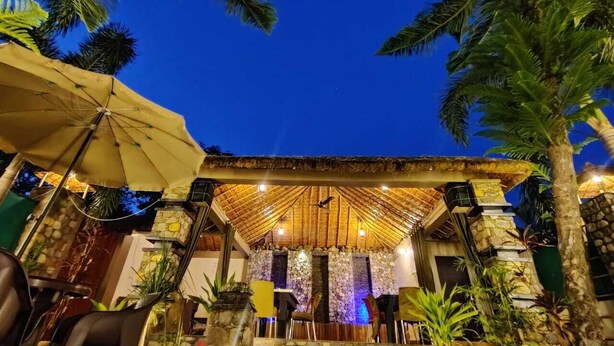 An outdoor dining area with a thatched roof, stone walls, and palm trees. - The Corbett Jungle  (Ramnagar)