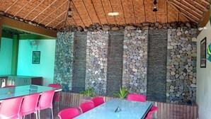 An indoor dining area with a thatched roof, stone wall accents, and a long table with chairs. - The Corbett Jungle  (Ramnagar)