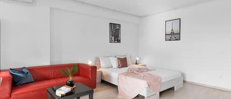 A modern, minimalist bedroom with a red sofa, a white bed, and a small table with a plant.