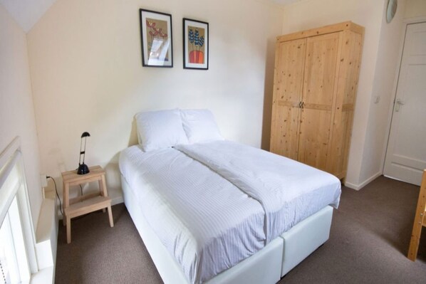 A bedroom with a bed, a wooden wardrobe, a bedside table with a lamp, and two framed pictures on the wall.