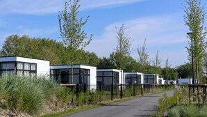 A row of modern, white, boxy buildings with large windows, surrounded by greenery and a paved path.