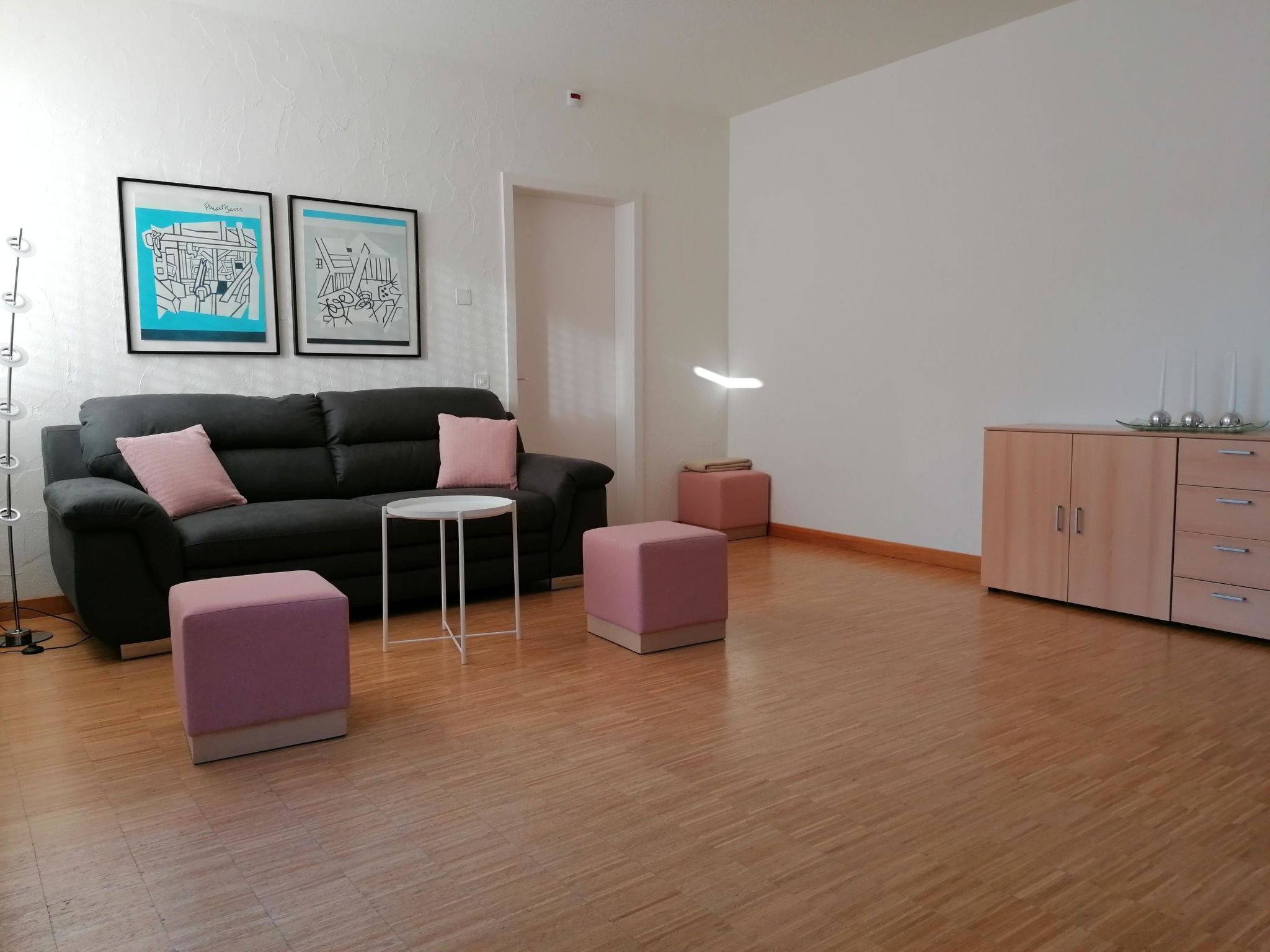 A modern living room with a black sofa, two pink ottomans, a small white table, and a wooden cabinet.