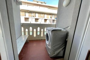 Ein Balkon mit Waschmaschine, einer Sitzbank und Blick auf ein Gebäude mit Balkonen.