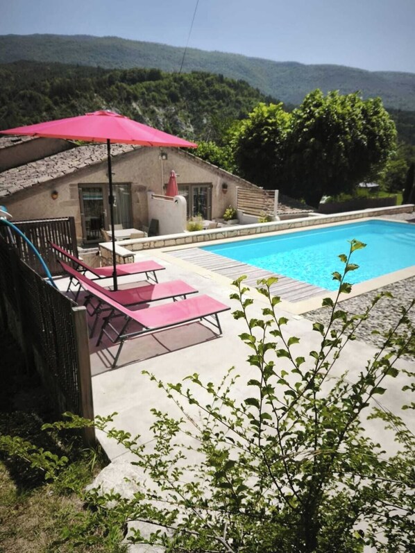 A swimming pool with lounge chairs and an umbrella, surrounded by a stone wall and greenery.