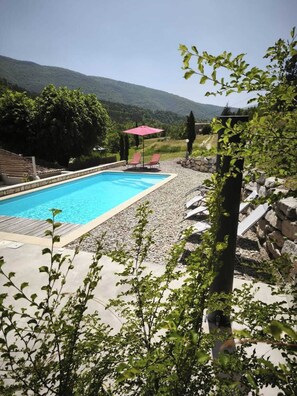 A swimming pool with lounge chairs and a red umbrella, surrounded by greenery and a stone wall.