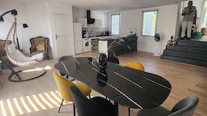 A modern kitchen with a central island, a dining area with a black table and yellow chairs, and a living area with a hanging chair and a staircase.