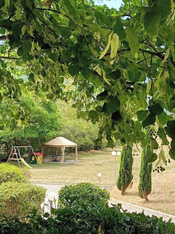 Un parque con zona de juegos, área de descanso cubierta y setos bien recortados.