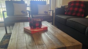 A living room with a wooden coffee table, two armchairs with moose pillows, a sofa with a red and black checkered pillow, and a view through large windows. - Lakeside 3 bed townhome w/ private beach access! (Windermere)