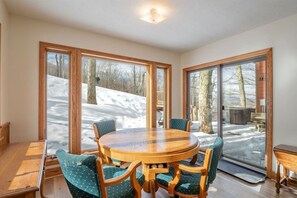 Ein Essbereich mit einem runden Holztisch und vier Stühlen, große Fenster mit Blick auf eine verschneite Landschaft und eine hölzerne Bank draußen.