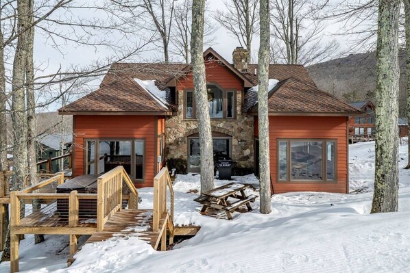 A two-story wooden cabin with a stone chimney, surrounded by snow and trees.