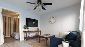 Ein modernes Wohnzimmer mit einer Couch, einem Couchtisch und einem Fernseher. Es gibt einen Deckenventilator, einen Spiegel und eine Pflanze.