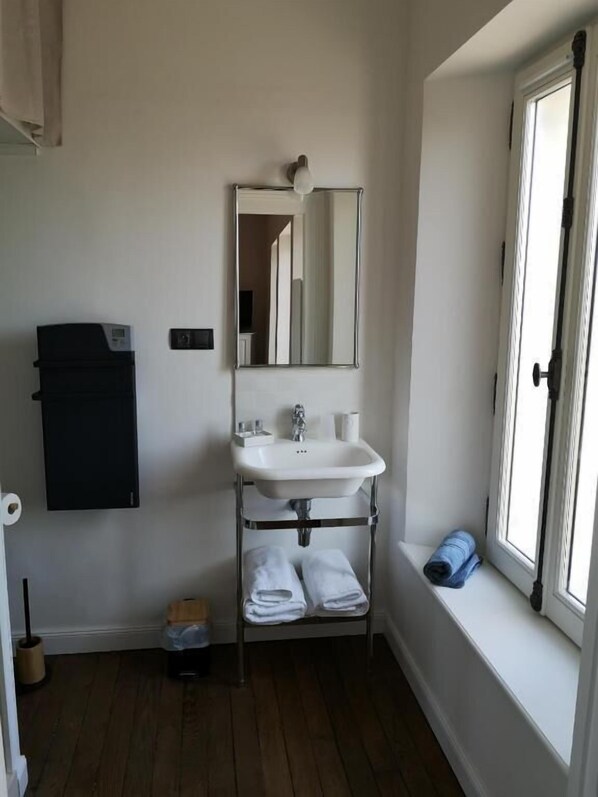 Un baño con un lavabo blanco, un espejo y una ventana. Hay un perchero con toallas y una toalla azul en el alféizar. Un armario negro está montado en la pared, y una escoba se ve en el suelo.