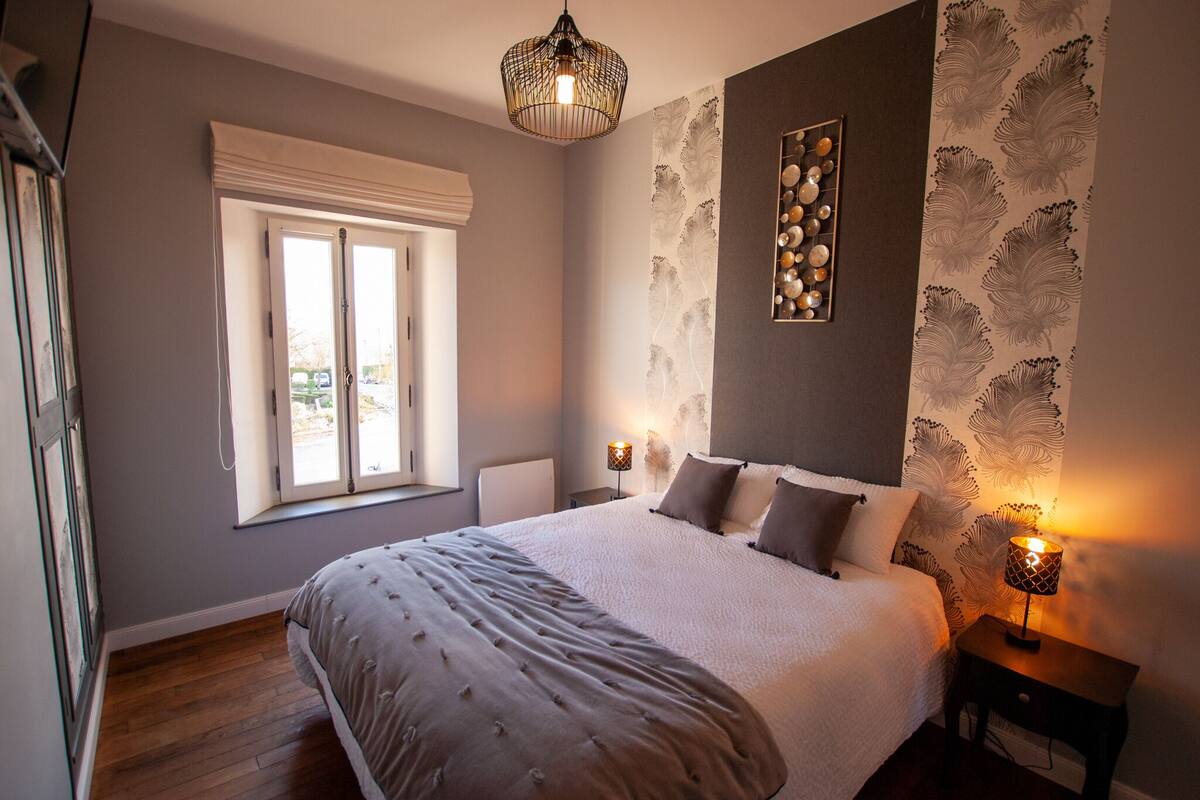 A bedroom with a large bed, a window with white blinds, a wall-mounted light fixture, and a decorative wall piece.