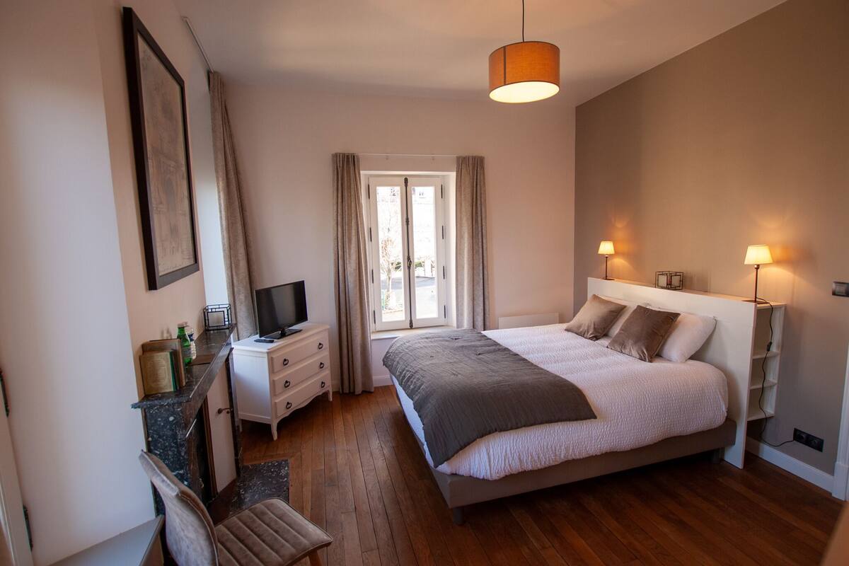 A bedroom with a bed, bedside lamps, a TV, a desk, and a window with curtains.
