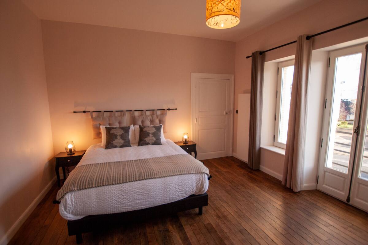 A bedroom with a bed, bedside lamps, a door, and a view through a window.