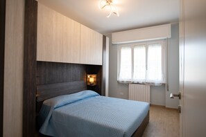 A bedroom with a bed, wooden wardrobe, bedside lamp, window with curtains, and a door. - LakeView (Polpenazze del Garda)