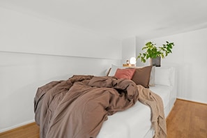 Cama bem arrumada com manta marrom, almofadas brancas e uma pequena mesa de cabeceira com abajur e planta.