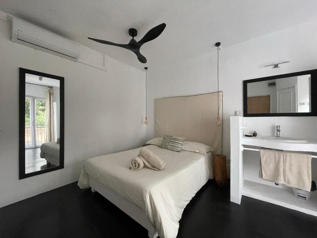 A bedroom with a bed, a ceiling fan, a mirror, and a bathroom with a sink and towel rack.