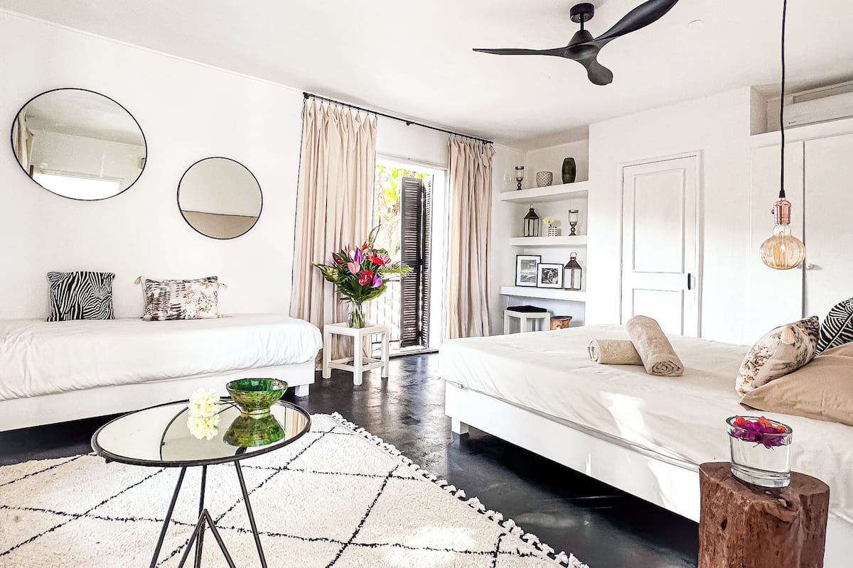 Une chambre moderne et bien éclairée, avec un grand lit, deux tables de chevet, une table basse ronde et un ventilateur de plafond.