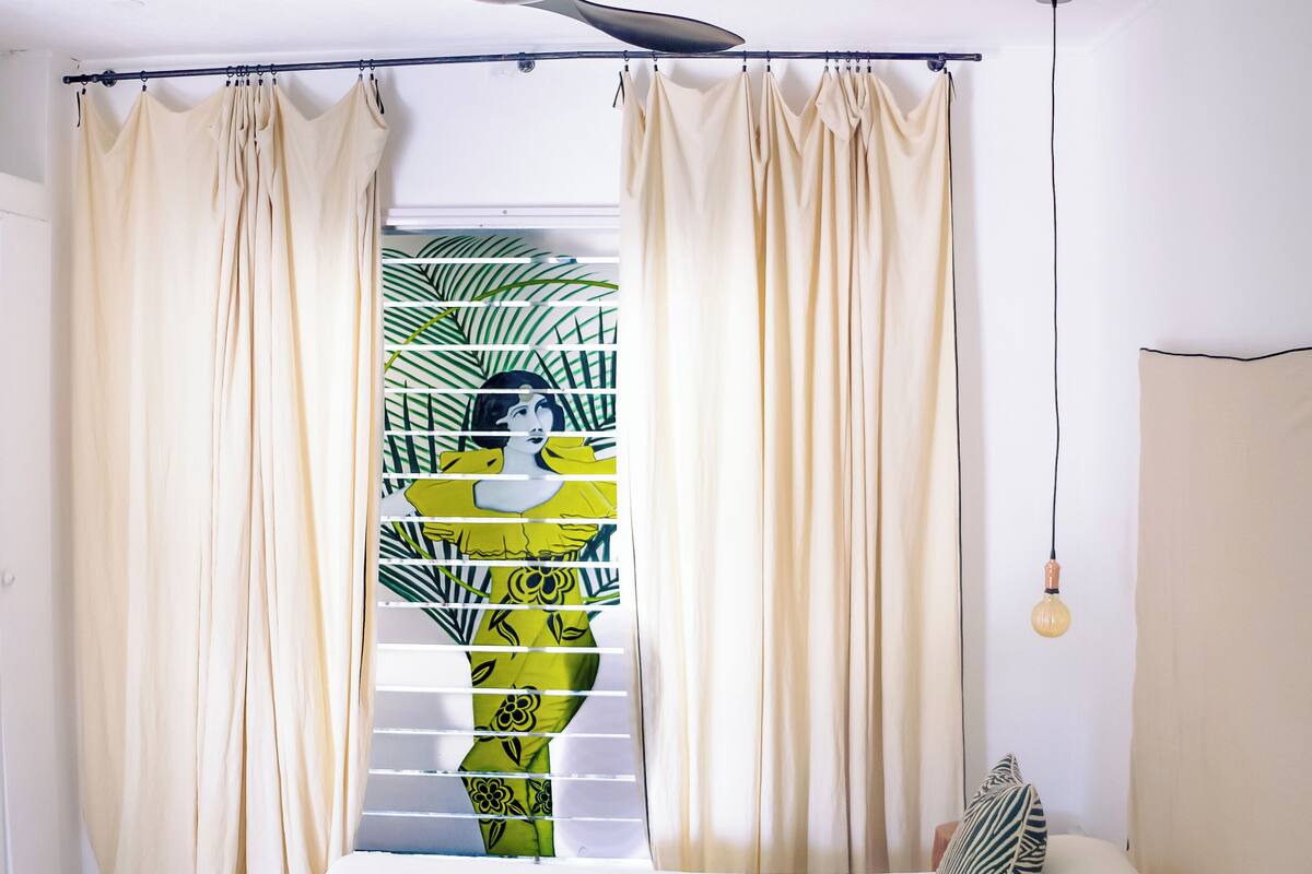 Une chambre à coucher avec un lit, des rideaux, un ventilateur de plafond et une vue sur les palmiers par la fenêtre.