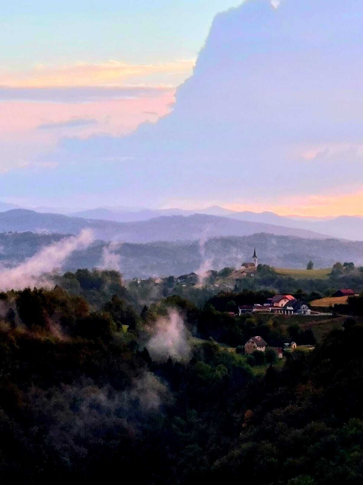 Una vista panoramica su un paesaggio rurale con una chiesa, case e fumo che sale dalle colline.