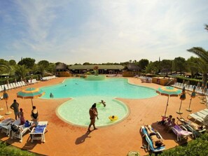 A large swimming pool with sun loungers and umbrellas, surrounded by palm trees and a building in the background.