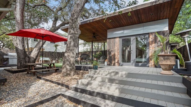 A covered patio area with steps leading to a modern building, surrounded by trees and outdoor furniture.