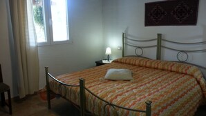 A bedroom with a metal-framed bed, a bedside table with a lamp, a window with curtains, and a wall-mounted decorative piece. - Agriturismo La Barca In Secca (Olmedo)