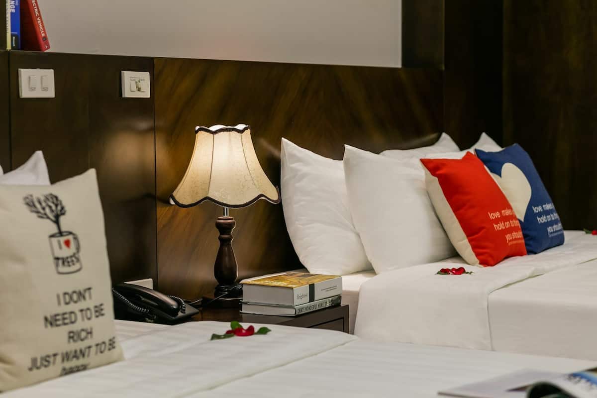 Quarto de hotel com cabeceira de madeira, abajur, telefone, um livro e almofadas decorativas.