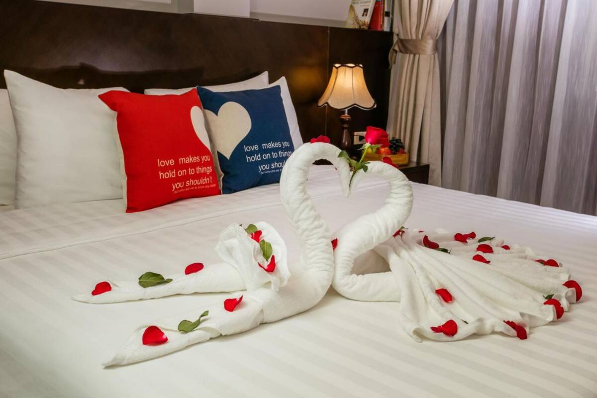 Cama com lençóis brancos, duas almofadas com desenhos de coração e um cisne decorativo feito de toalhas e pétalas de rosa.