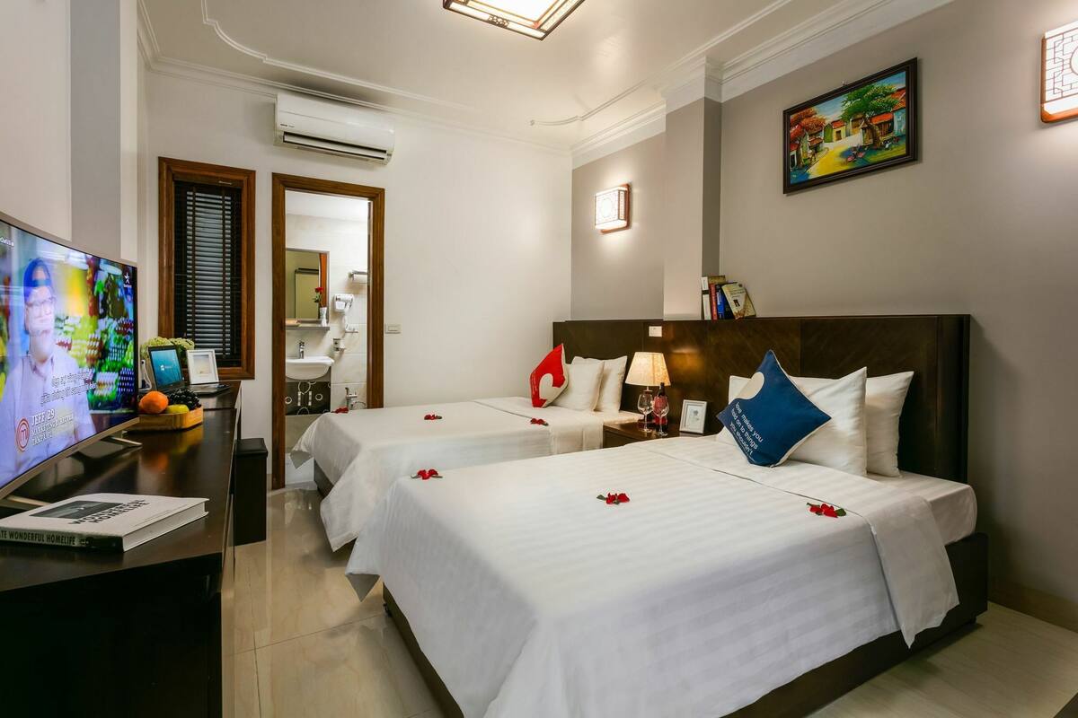 Quarto de hotel com duas camas, uma mesa de trabalho e televisão, e um banheiro visível através de uma porta.